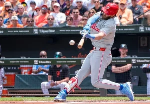 A Philadelphia Phillies baseball player in a gray away uniform with red trim and blue cleats swings at a pitch during a game, with the baseball visible near his bat. The player wears a red cap with the Phillies 'P' logo. A crowd of spectators fills the stadium stands in the background.