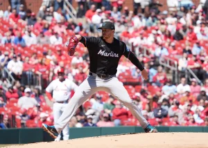 A left-handed baseball pitcher in mid-delivery on the pitcher's mound during a professional baseball game. The pitcher wears a black Miami Marlins jersey with "Marlins" written in white script across the chest, a black cap with the Marlins logo, white pants, and bright blue cleats. He's captured in his pitching motion with his right leg extended and left arm drawn back preparing to release the ball, which is visible in his gloved hand. The background shows a packed stadium with distinctive red seats filled with spectators. The setting appears to be a sunny day game, with the pitcher casting a shadow on the dirt mound. A blurred player in white can be seen in the background on the field.
